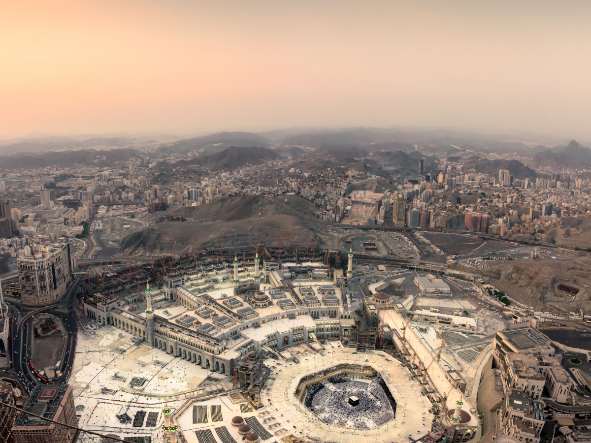 Aerial view of the Kaaba in Mecca, surrounded by a multitude of pilgrims in white garments, with the cityscape in the background.