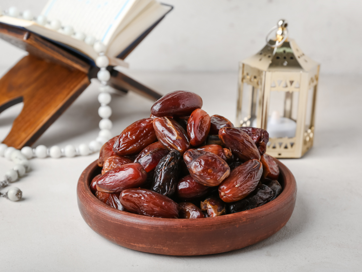 A bowl of ripe dates placed next to a Quran and prayer beads on a light surface, with a lantern in the background