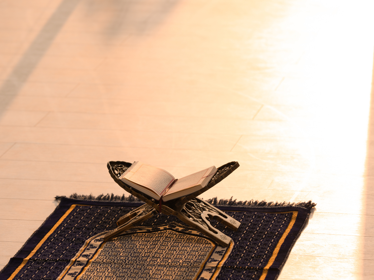 An open Quran book resting on an ornate prayer mat, illuminated by sunlight, emphasizing the sacredness and tranquility of the setting.