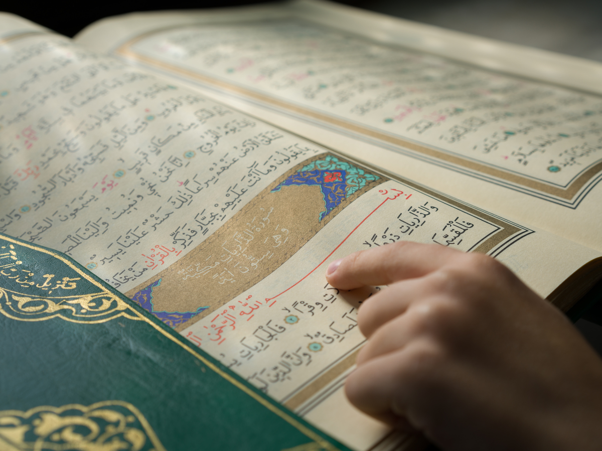 A person's hand pointing to a verse in an open Quran, highlighted by the intricate detail and colorful text of the scripture.